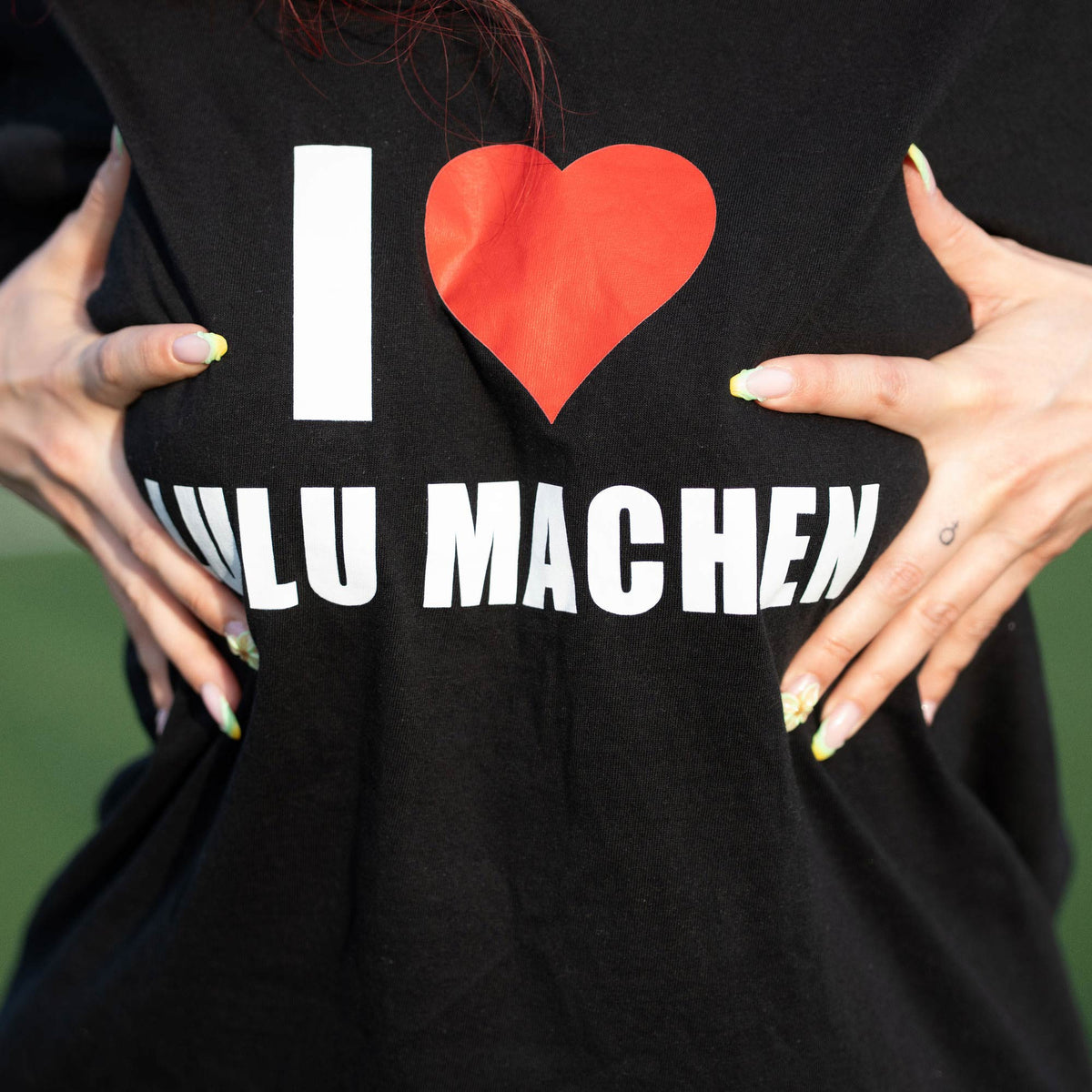 Nahaufnahme des schwarzen T-Shirts, Model hält Frontprint „I Love Lulu Machen" mit rotem Herz ins Bild.