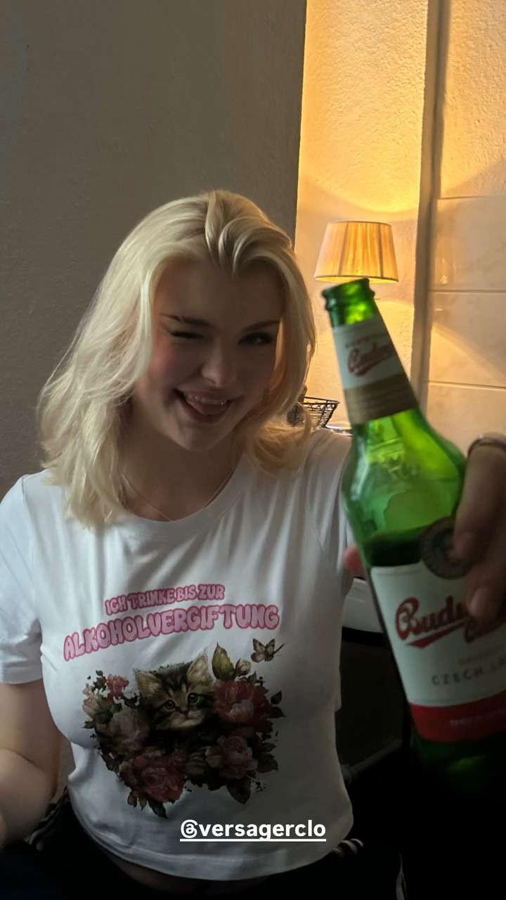 Junge Frau mit blondem Haar trägt ein T-Shirt mit Katzenmotiv und Aufdruck „Ich trinke bis zur Alkoholvergiftung“ und hält eine Bierflasche in die Kamera