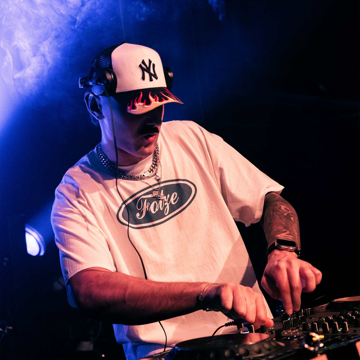 Model trägt weißes „Fotze"-T-Shirt mit Oval-Frontprint am DJ-Pult, blaues Clublicht im Hintergrund