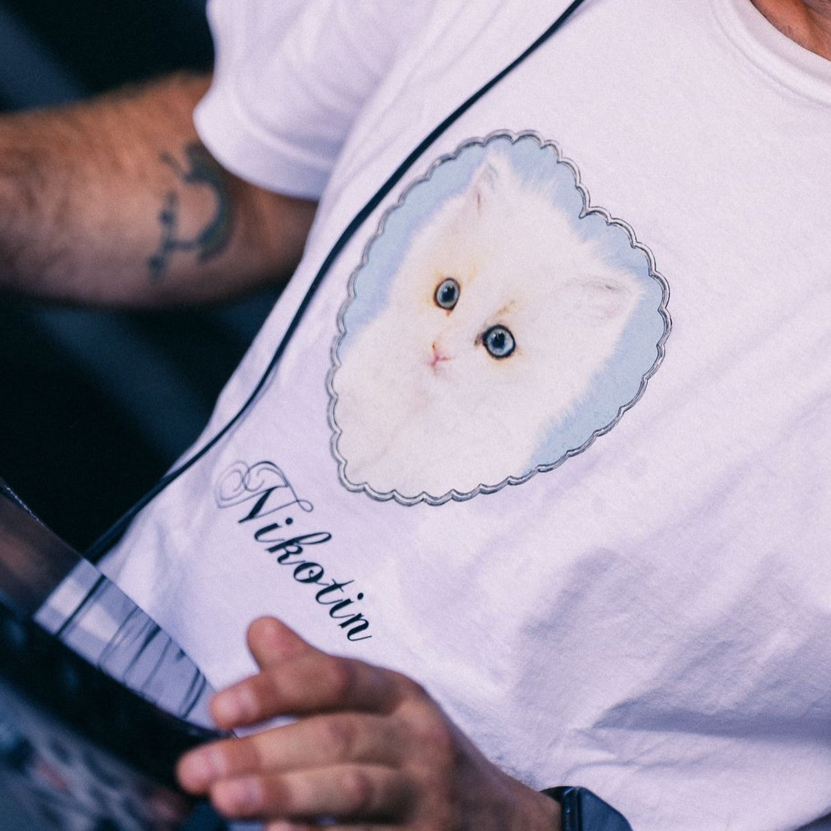 Nahaufnahme: Model mit Tattoos trägt weißes T-Shirt, Frontprint zeigt Kätzchen im Herzrahmen mit Schriftzug „Nikotin"